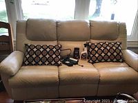 Full front view of the three-seat beige leather electric reclining sofa with pillows and accessories on seat.