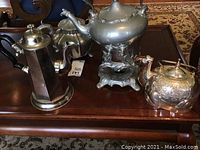 Four antique metal coffee and tea pots displayed on a wooden table, showing different shapes and designs including the camel-shaped silver plated pot.