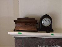 Tea caddy and mantel clock shown side by side on a white shelf above a stone fireplace.
