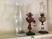 View of the three oil lamps and the large clear glass hurricane globe. The porcelain oil lamp has a pink floral decorated base. Small pottery oil light in the front.