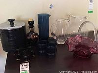 Lot displayed on a black tabletop including cranberry glass basket, clear and colored pitchers, decanter, shot glasses, and vintage ice bucket.
