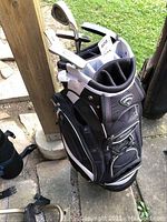 Full view of black and gray Callaway golf bag filled with assorted golf clubs standing upright outdoors on stone floor.