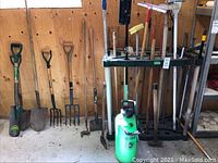 Wide view of tool rack holding the yard tools including shovels, pitchfork, hoes, brooms, an axe, and a garden sprayer on the floor.