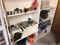 Shelving unit with multiple garden hand tools including chisels, scrapers, trimmers, birdhouse on the right shelf, orange and blue buckets below