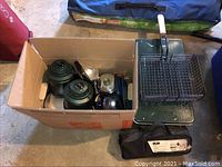 Photo showing a box containing green enamel camping pots, pans, kettle, and Coleman lantern flashlights. Coleman propane camp stove with grill basket placed beside the box.