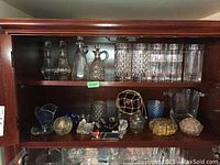 Full view of shelf containing vintage glassware, crystal cruets, tumblers, paperweights, and decorative glass items.