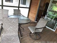 Full view of patio set on wooden deck showing round glass top table and four swivel chairs with fabric seats and backs in leaf pattern.