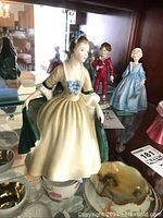Full front and side view of the Royal Doulton figurine in cream dress with green cape and blue bow, shown on glass shelf with ceramic bowls and smaller porcelain figures in background.