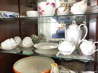 Glass cabinet shelf holding the tea set items including cups, saucers, sugar bowl, creamer, teapot, and plate.