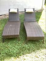 Photo of two resin wicker outdoor loungers placed side-by-side on grass with cream-colored pillows attached to the headrests.