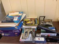 Photo shows a lot of office supplies including mailing labels, legal size file folders, a stapler, and a stapling gun in original packaging on a wooden surface.