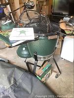 Full view of green ceramic grill on stand with lid open, showing cookbooks on side shelf and cover on floor
