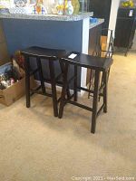 Pair of black wood bar stools standing side by side on carpet near kitchen counter, showing overall condition and shape