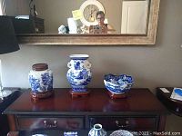 Photo of three blue and white pottery items with wooden stands on a dark wood console under a large wall mirror.
