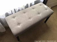 Straight angled view of grey fabric button tufted boudoir bench with black legs on carpet flooring near window curtains