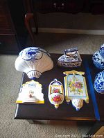 Overhead photo showing all ceramic and painted iron items in the lot on a dark table surface, including blue and white planter, floral wall plaques, and small painted iron piece with handle.