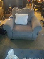 Front view of a large cushioned chair with rounded armrests and a soft pillow. Beige/light grey fabric upholstery showing some wear.