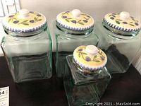 Front view of all four canisters showing the square shape, clear heavy glass material, and ceramic lids with hand-painted blue berry and leaf design.