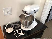 White KitchenAid Classic stand mixer on black cabinet. Stainless steel bowl attached. Flat beater, wire whisk and dough hook attachments displayed next to mixer.
