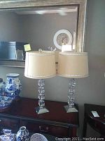 Pair of matching table lamps on sideboard with mirror behind. Each lamp has stacked glass cube bases and beige drum shades with decorative trim.