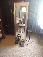 Photo showing full tall mirror with silver-toned plastic frame leaning against the wall. Black metal candle holder, small black lantern, and metal hook placed in front on carpeted floor. Curtain and furniture visible in background.