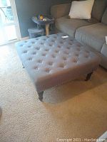 Large square tufted gray fabric footrest or sofa table with wooden legs and wheels placed near sofa on carpeted floor.