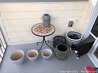 Patio table with mosaic tile top on wrought iron base, ceramic plant pots, candle lantern, and woven baskets on porch floor, cat visible.