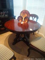 Round solid wood dining table with pedestal base seen from front angle, showing table surface and base, surrounded by chairs (not included).