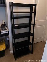 Front view of tall black collapsible bookcase with five shelves against wall next to door and table.
