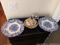 Photo of two blue and white ceramic serving plates with floral patterns and a yellow cheese dish with lid, plus a napkin holder, displayed on a black surface against a white wall.