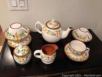 Full tea set displayed on black surface showing teapot, creamer, sugar bowl, cups, and saucers with floral decoration and terracotta rim.