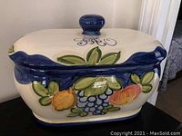 Front and side view of the ceramic bread box showing painted fruits and leaves, blue trim, and lid knob.