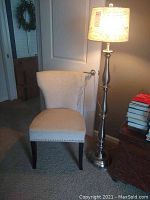 Cloth covered beige side chair with nail brads and frayed fabric on back, next to stainless steel floor lamp with beige shade, lamp turned on showing light output.