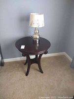Round wood side table with lamp on top against gray wall on carpeted floor.