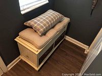 Side view of the white wood composite storage bench with cushions on top and two wicker baskets underneath placed in a corner between two dark-colored walls on wood flooring.