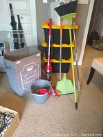 Dirt Devil stick vacuum, two Black & Decker cordless dust busters, grey laundry basket labeled 'Deluxe Laundry Service', green broom, dustpan, mop bucket with cleaning tools.