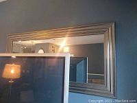 Front view of large rectangular wall mirror with silver-colored frame showing beveled edges on glass and reflection of the room.