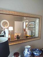 Front view of large rectangular wall mirror reflecting a room with lamps and decorative items, showing the gold frame and beveled glass edges.
