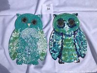 Two teal colored owl plaques displayed side by side on a white fabric background, showing front details.