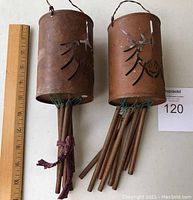 Two cylindrical rustic metal wind chimes with leaf cut-out designs and metal hanging tubes, shown next to a ruler for scale.