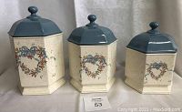 Front view of the three hexagonal ceramic canisters with blue lids displaying the heart-shaped floral wreath decoration and geese illustrations