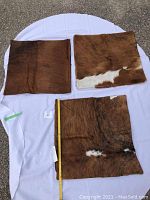 Three large square cowhide pillow covers laid flat on white cloth with measuring tape showing approximate size.