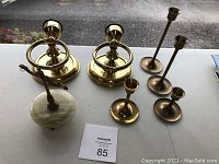 All items on a white flat surface including 2 brass wall sconces, 4 freestanding brass candle holders, and one brass and stone decorative item.