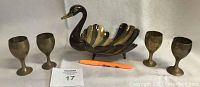 Photo of a brass swan sculpture with curved neck and layered feather wings and four brass goblets arranged around it on a grey surface, showcasing entire lot.