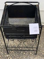 Front angled view of a black metal flower pot stand showing the square basin, circular top ring, decorative scroll details on the supporting frame, and rust spots on edges and legs.