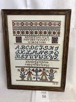 Full view of framed sampler showing entire design including alphabets, numbers, floral and geometric motifs, figure, and signature dated 1991.