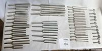 Large assortment of tuning forks arranged in rows on fabric, showing a range of sizes and handle shapes; all metallic silver metal color.