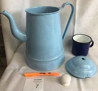 Front view of blue metal teapot with lid and dark blue enamel cup on white cloth background