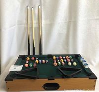 Miniature pool table with accessories arranged, three cues standing upright behind, four triangle racks on table, pool balls laid out on table, chalk cubes placed on corner.