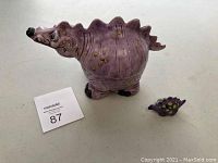 Pink ceramic dinosaur figure with detailed textured body and painted yellow spots, next to a small purple dinosaur pencil sharpener with yellow spots on a white surface.